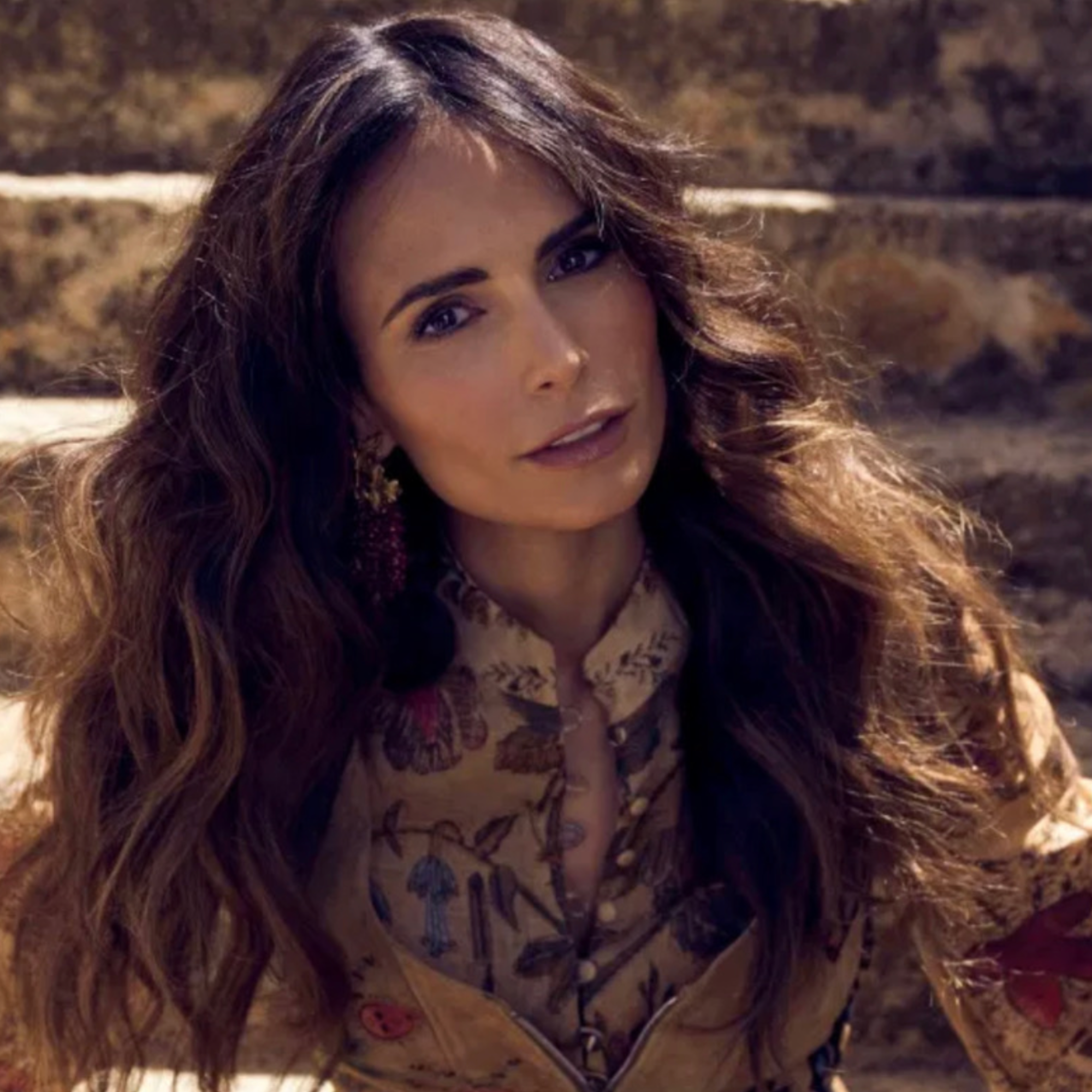 Woman with long wavy hair wearing a patterned blouse outdoors.