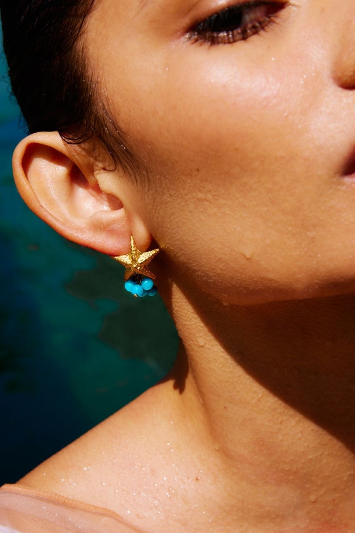 Close-up of a person wearing gold earrings with turquoise stones against a blurred natural background.