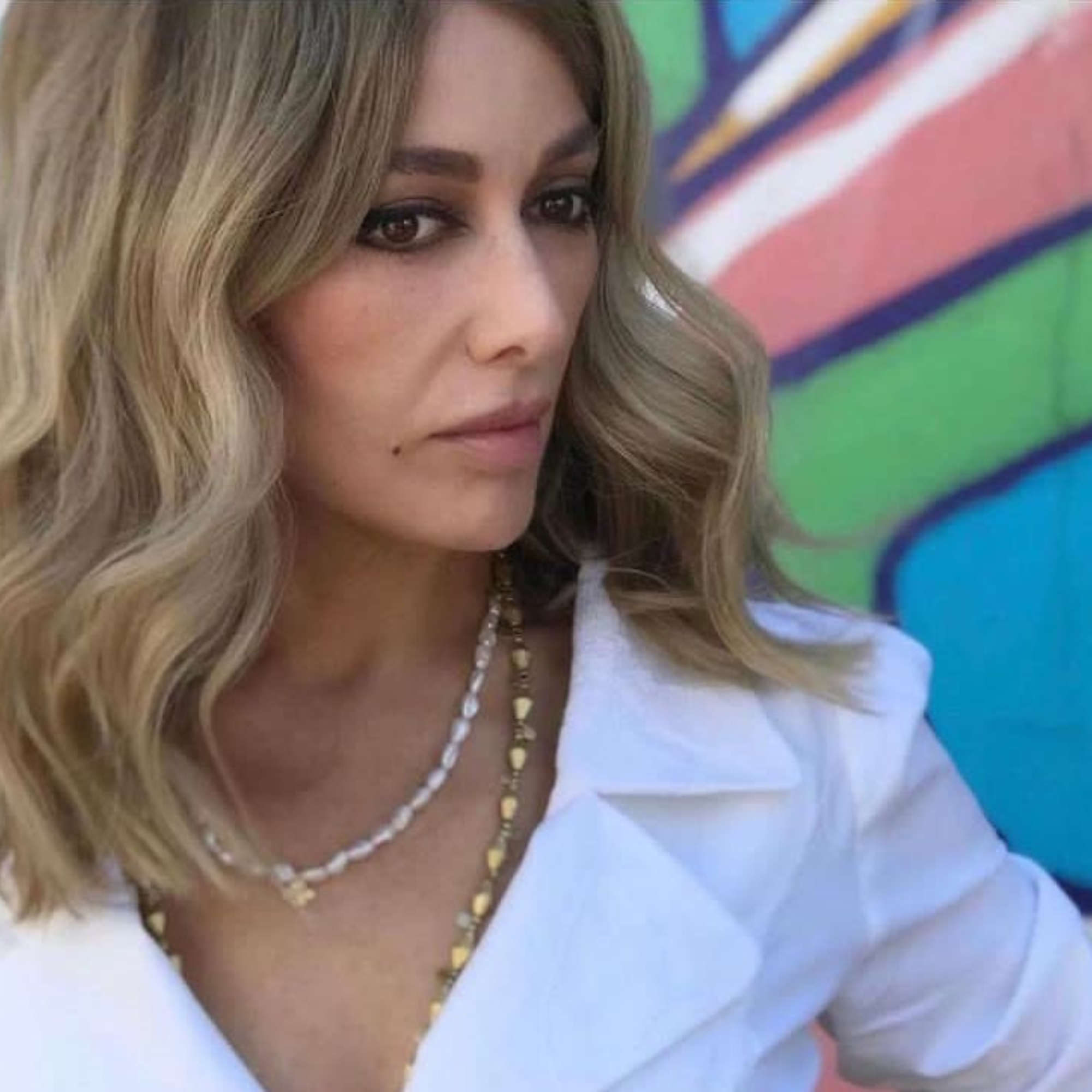 Woman with blonde hair wearing a white outfit and gold necklace against a colorful background