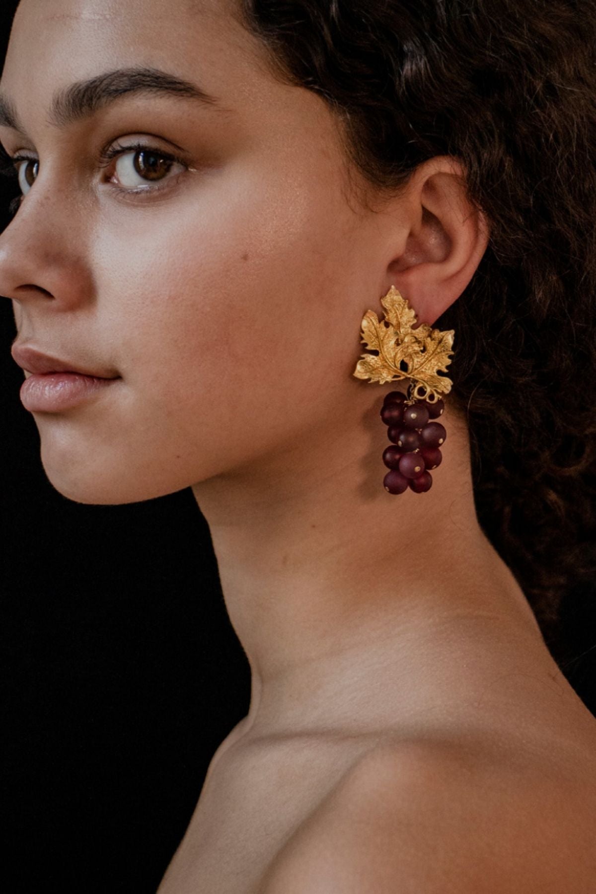 Close-up of a woman wearing gold earrings with grape designs against a black background