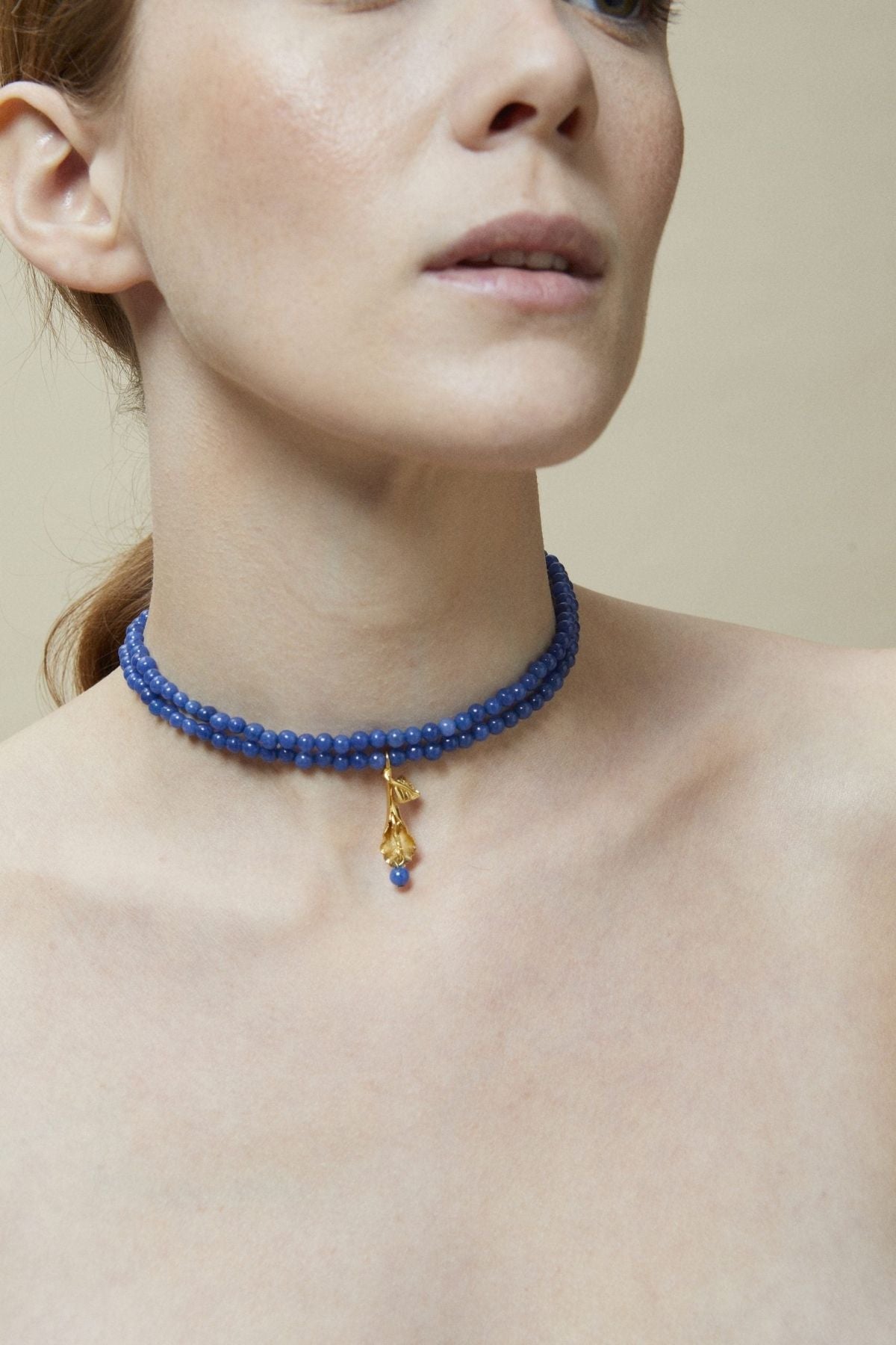 Woman wearing a blue beaded choker necklace with a gold pendant on a neutral background