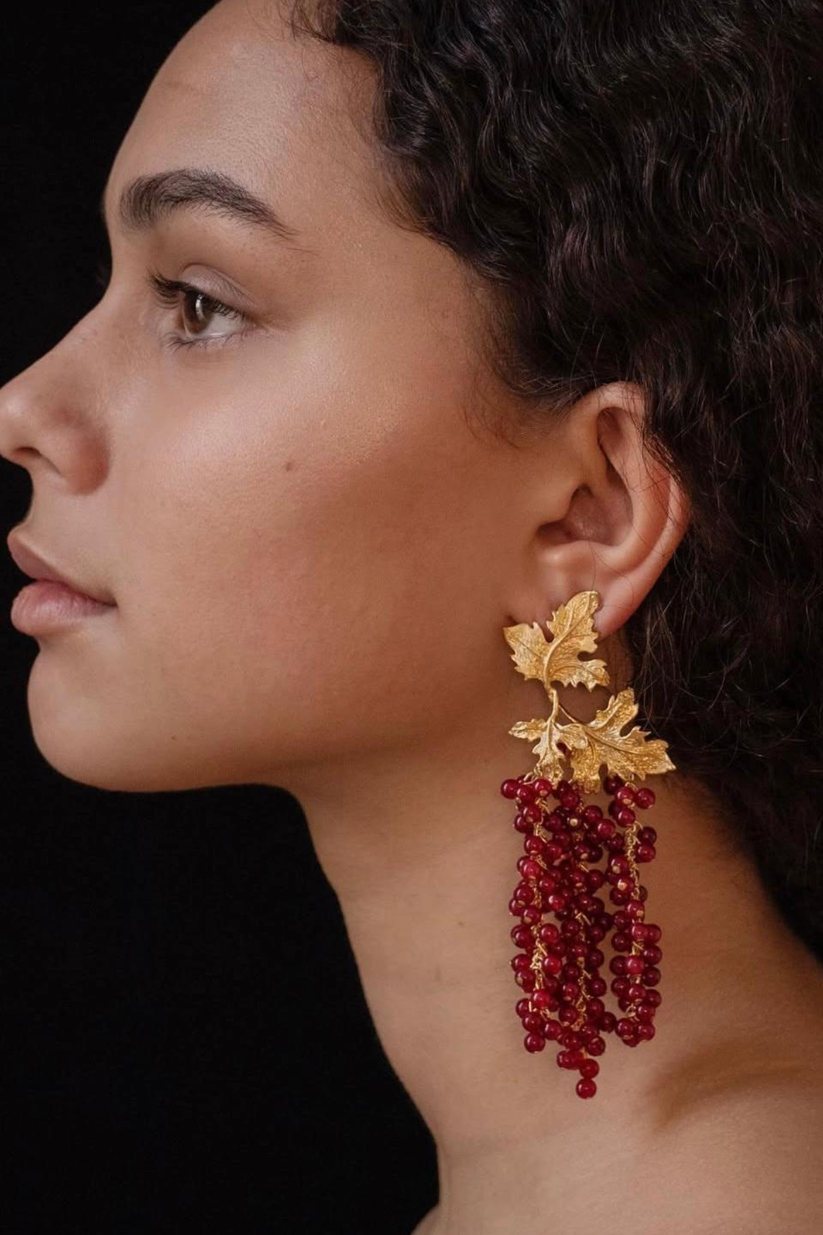 Close-up of a person wearing gold leaf and red bead earrings against a black background