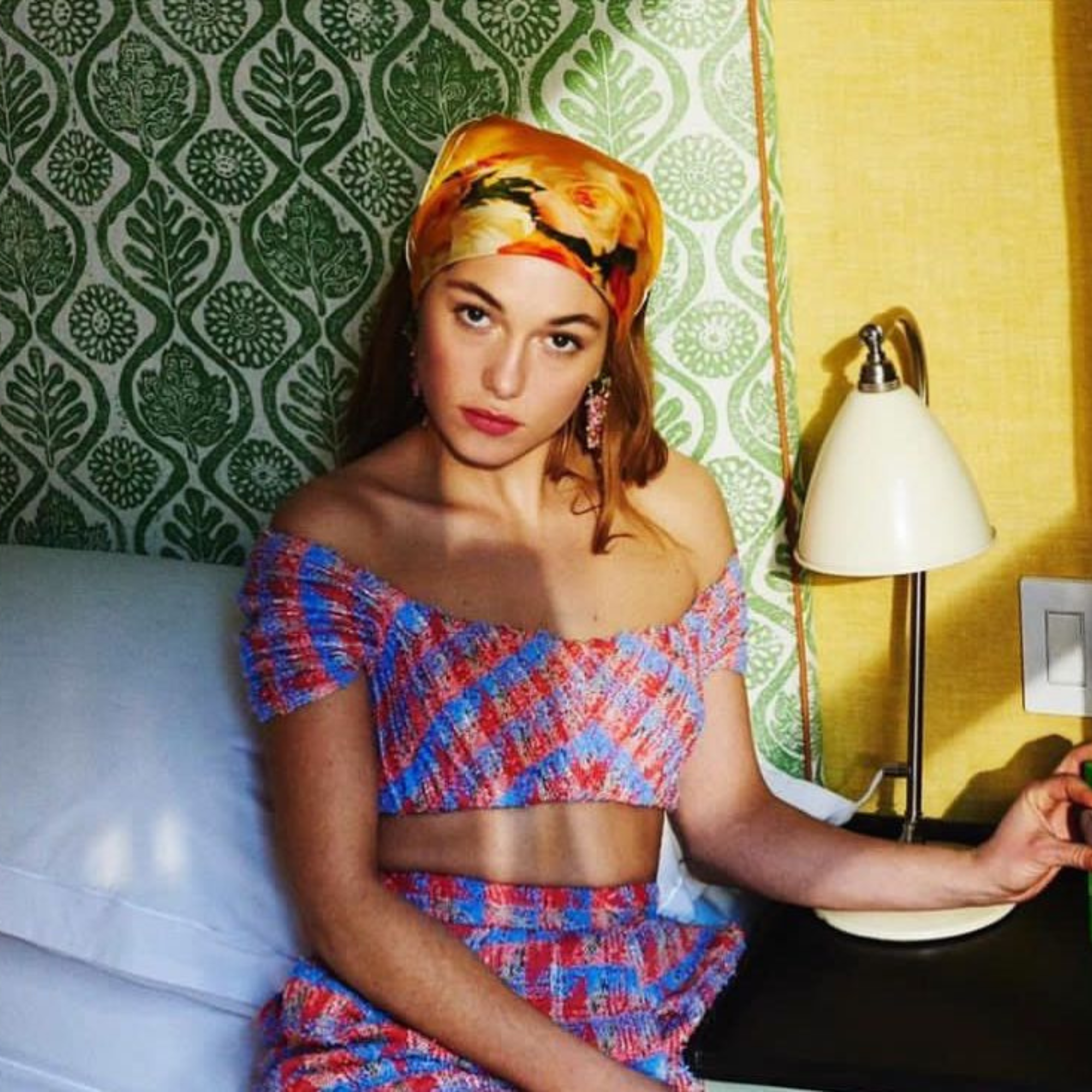 Woman in a colorful outfit sitting on a bed with patterned wall and lamp in the background