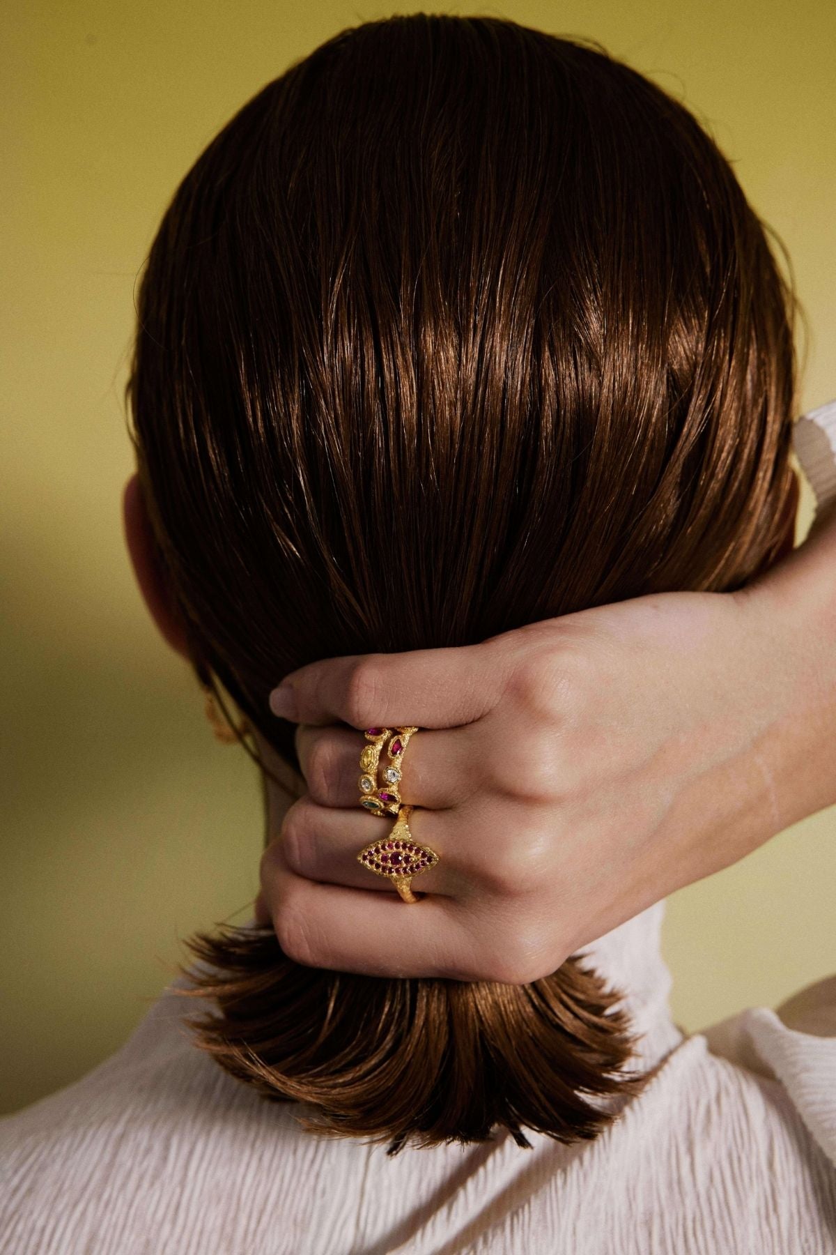 Person with a bun and hand adjusting it, wearing a gold ring with a red gemstone on a yellow background